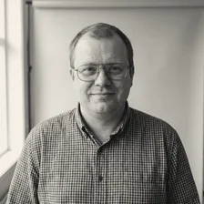 Jørgen Solheim smiler svakt mot kameraet, med briller og rutete skjorte i blått og brunt, foran en lys vegg ved et vindu.