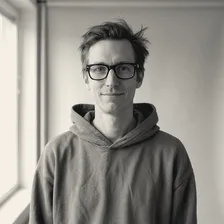 Mathias Lindqvist med bustete hår, svarte briller og grå hettegenser, smiler lett mot kameraet foran en hvit vegg ved et vindu.