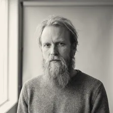 Trond Hagen ser alvorlig inn i kameraet, med langt lyst hår, stort skjegg og grå strikkegenser, foran en lys vegg ved et vindu.